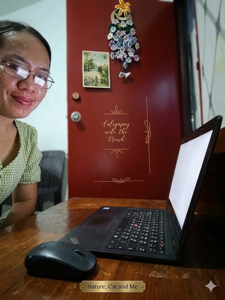 An angled, medium shot of a content creator sitting at a rustic wooden desk, viewed from the side. On the desk sits a sleek black laptop with a completely blank, glowing white screen, a black wireless mouse, and a small gold nameplate at the bottom edge that reads "Nature, Cat and Me." The creator, a woman with dark hair and glasses wearing a green and white checkered blouse, smiles toward the camera from the left side of the frame. In the background, a dark red door is decorated with an intricate hanging arrangement of white and blue artificial flowers and a small landscape picture postcard. Fine gold line art and elegant cursive text are overlaid on the center of the door.